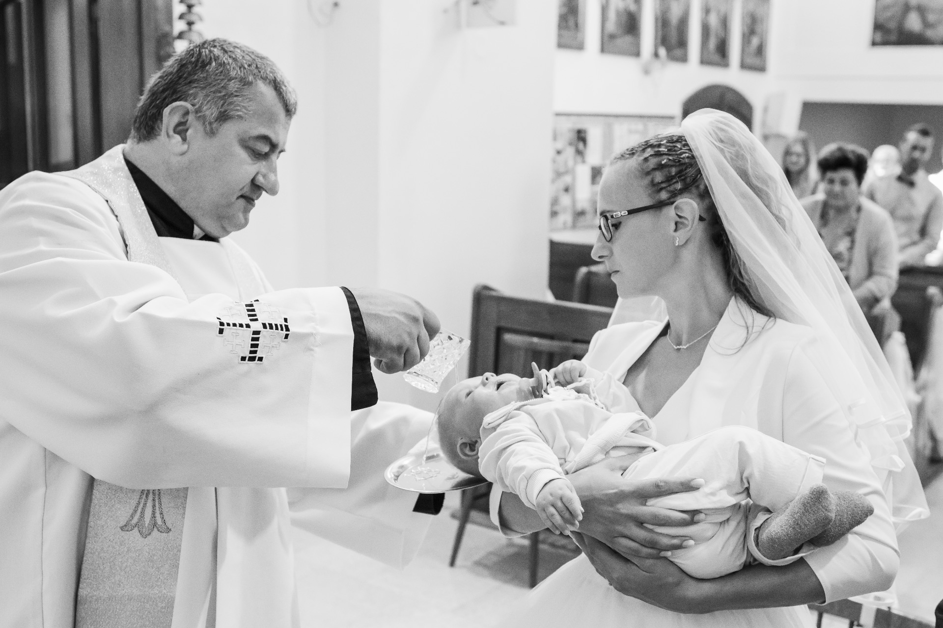 Kněz v bílém rouchu drží dítě, matka v závoji se sklání, v pozadí lidé v kostele.