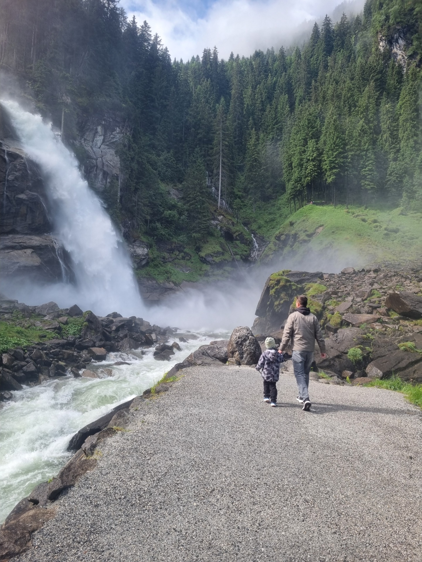 Turista stojí na skalní cestě u vodopádu, mlha stoupá, v pozadí hustý horský les.